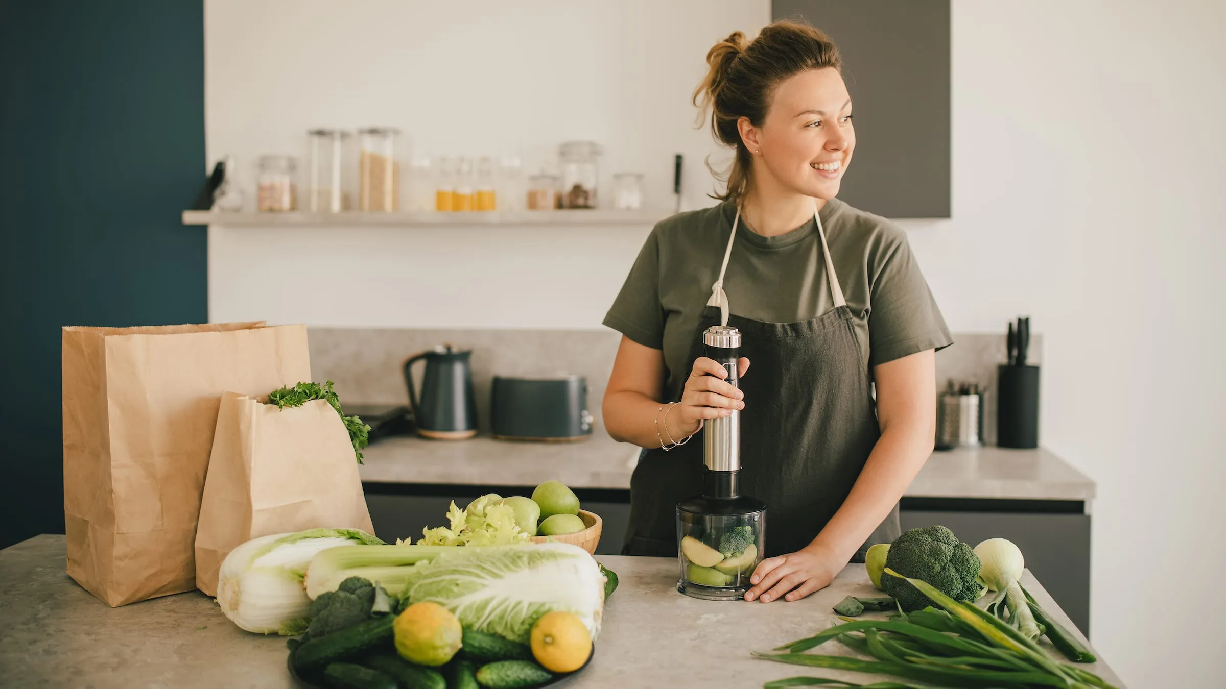 Young woman making smoothie at the kitchen, using blender and fresh fruits and vegetables.